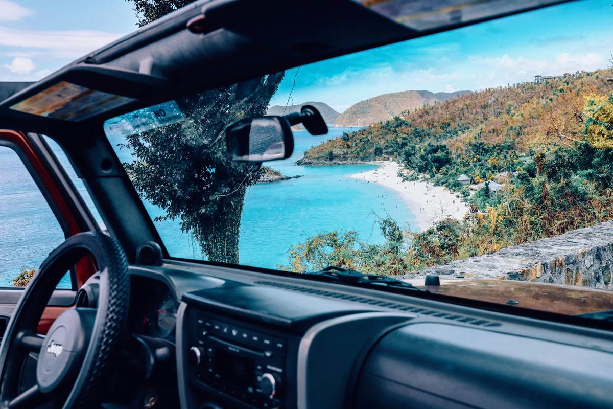 View from inside a car driving along St. John's coastal road with pristine beach and turquoise waters visible through the windshield